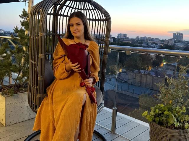 Uma mulher com cabelo castanho, vestido amarelo e lenço vermelho, sentada em uma cadeira de vime em um terraço, segurando uma bolsa marrom, com o pôr do sol ao fundo e a silhueta da cidade visível..