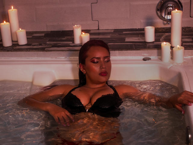 LaurenMartins in a black bralette, floating in a bathtub with candles, water up to her tits, red lipstick, dark wet hair, relaxed, arms resting on tub edges, eyes closed.