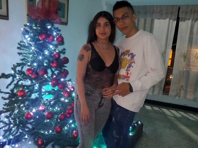 ZafiroModels, a woman with tits in a sheer black top and gray pants, stands next to a man in a white sweatshirt, by a blue-lit Christmas tree.