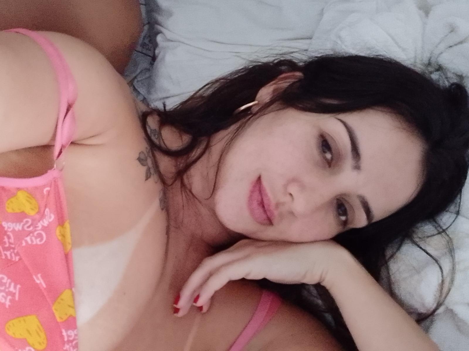 HotGiselle69 com cabelo escuro, blusa rosa, estampas amarelas, deitada na cama, cabeça apoiada na mão, sorriso discreto, pequena tatuagem no peito, alças de sutiã rosa, esmalte rosa, relaxada..