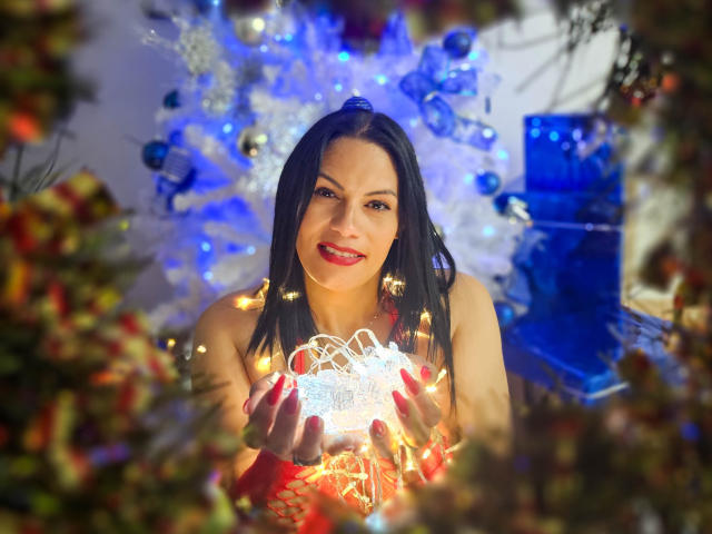 LizaRussell with black hair, red lipstick, and red fishnet gloves, holding glowing white ornament, standing in front of blue and white Christmas tree, surrounded by blurred Christmas decorations.