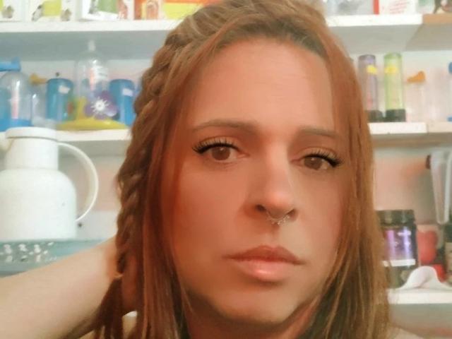 RubyReal has long brown hair, a nose ring, and is pouting with slightly parted lips. Her makeup includes long eyelashes. Shelves with various items are in the background.