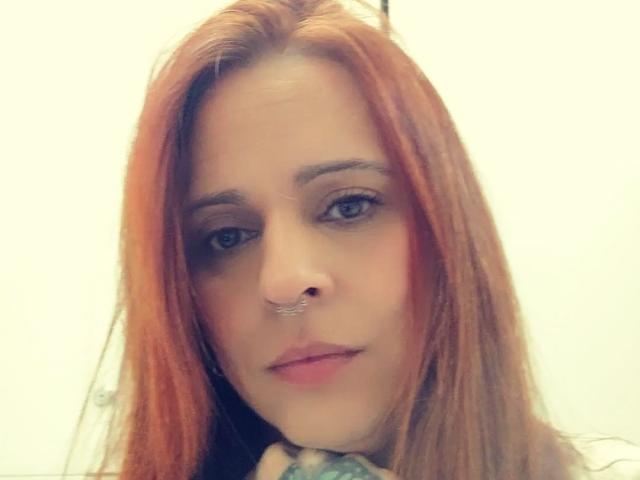 RubyReal has red hair, a nose ring, and a slight smile. Her blue eyes are looking at the camera. She's touching her chin with a tattooed hand.