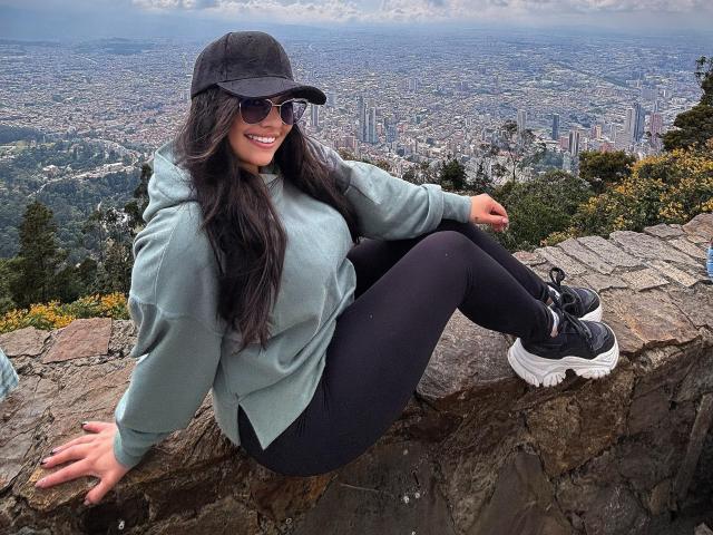BellaSmithX69 with long dark hair, wearing a black cap, sunglasses, green hoodie, black pants, and black sneakers, sitting on a rocky ledge, smiling, showing her ass.