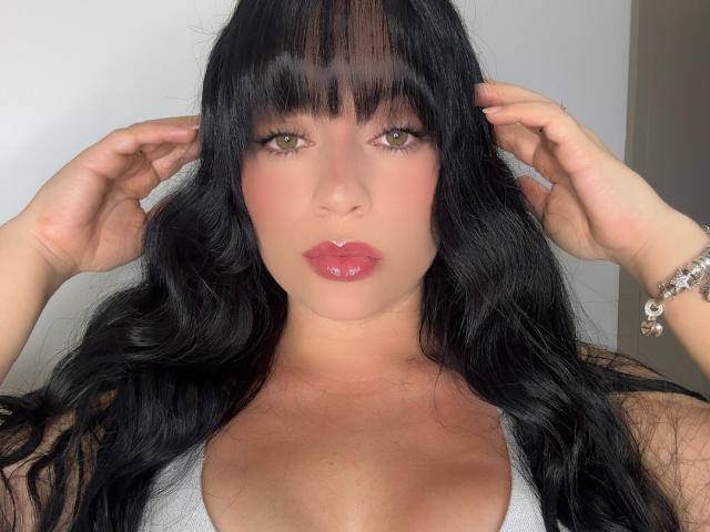 PammelaPrice has long black hair, green eyes, pink lips, and is wearing a white tank top. She's touching her hair with both hands, showing a silver bracelet.