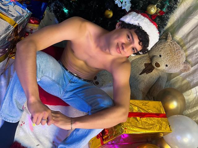 Shirtless SamuelHills with curly dark hair, wearing a Santa hat, sits on red and white cloth, blue jeans, and black socks, beside a large teddy bear and Christmas gifts.