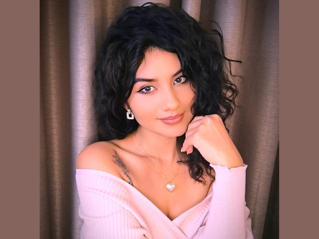 CanelaLebrad with curly black hair, pink off-shoulder top, heart necklace, tattoos on right shoulder, earrings, and pink lipstick, smiling softly, hand on cheek, brown curtain background.