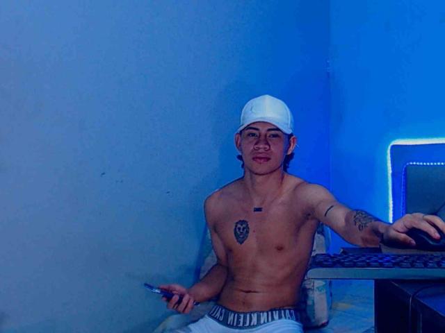 Logannth is shirtless, wearing a white cap, holding a pen, sitting at a desk with a laptop, tattoos on chest and arm, blue room, gray pants, smiling.