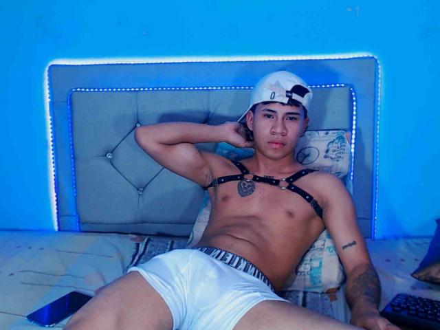 Logannth, shirtless, wearing white boxer briefs, black harness, white cap, laying on bed, arm behind head, blue light, visible tattoo on right arm, and a flaccid, uncircumcised, small penis.