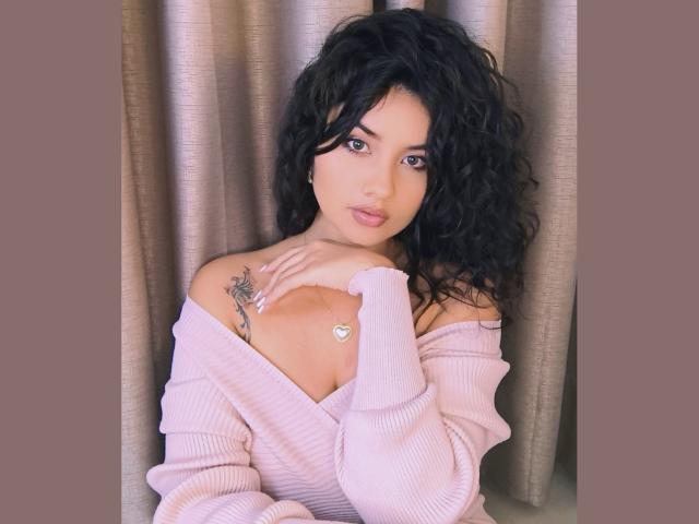 CanelaLebrad with curly black hair, pink off-shoulder sweater, heart necklace, tattoo on right shoulder, looking at camera, hand on collarbone, brown curtains in background.
