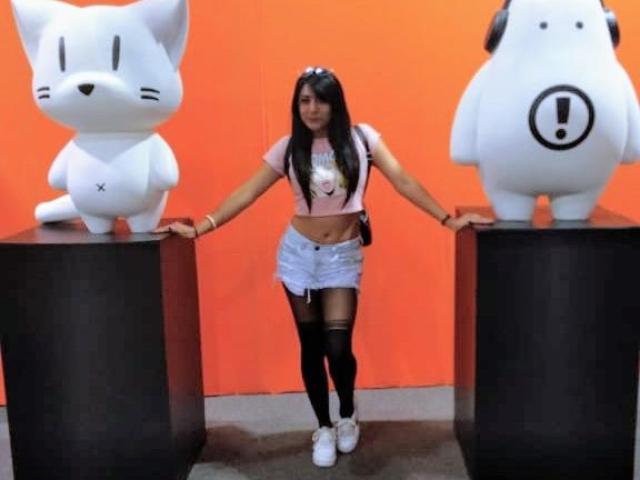 SophiQueen with long black hair, wearing a pink crop top, white shorts, black stockings, and white sneakers, stands between two white plush toys, one with an X and the other with an!.