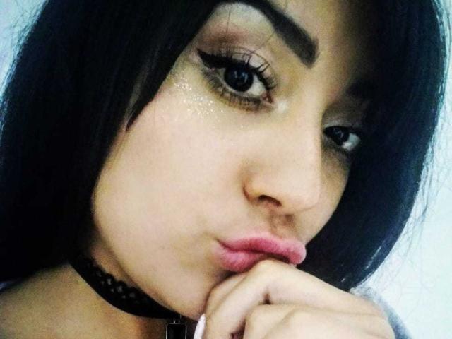 SophiQueen with dark hair, thick eyebrows, glossy pink lips, and a black choker, pouting and biting her finger, showing off her sexy, shiny eyes and soft skin.