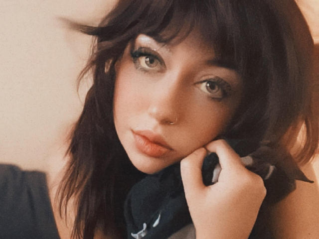 MookieMay has dark brown, wavy hair, green eyes, and a small nose ring. She's holding headphones near her chin with a serious, slightly pouty expression.
