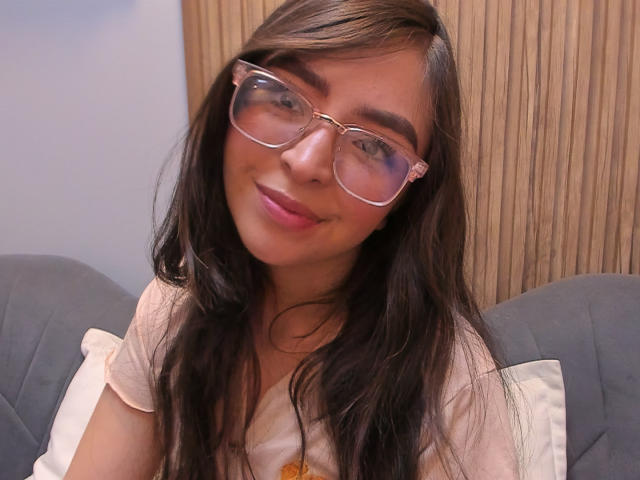 EvaMcAllister with long brown hair, wearing large clear glasses and a pink shirt, smiling softly at the camera, showing a cute face with pink lipstick and subtle makeup.