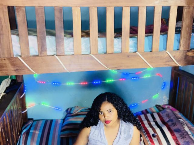 RineeAimee with curly black hair and red lipstick, wearing a grey sleeveless top, sitting below a wooden bunk bed with colorful Christmas lights, on a striped bed.