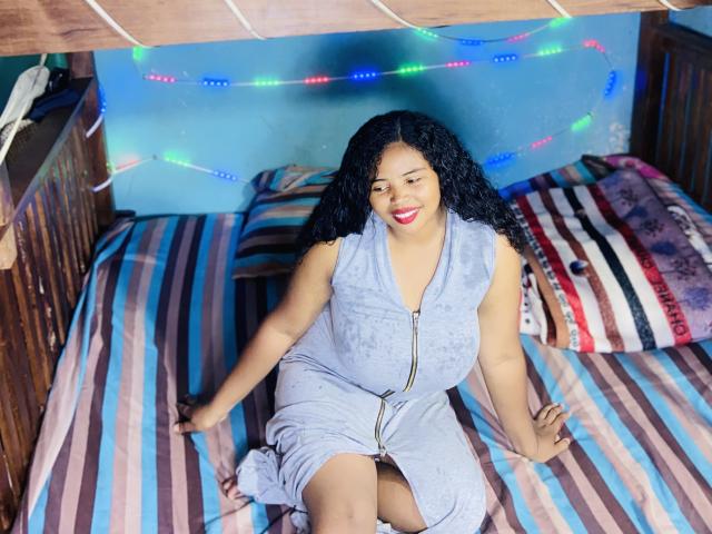 RineeAimee with curly black hair, medium brown skin, red lipstick, and a gray zip-up dress, sitting on a striped bed with colorful string lights above.