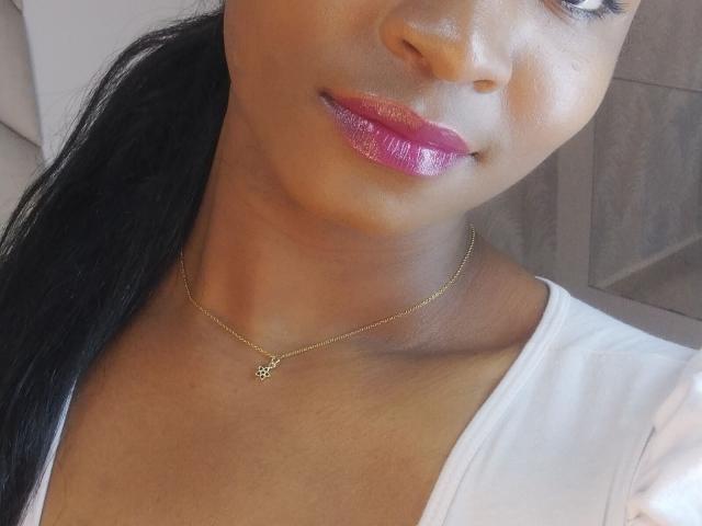 CaritoPerez has glossy pink lipstick, a gold necklace with a small charm, and a white shirt, with dark hair in a ponytail. Her skin is smooth and brown.