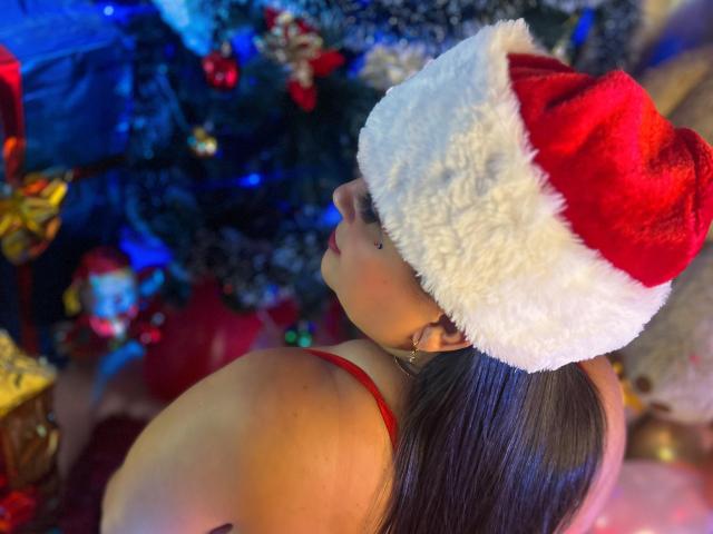 KerlyDolly with long brown hair, wearing a red Santa hat and red bra, looking over her shoulder, showing a hint of her tits, by a decorated Christmas tree.