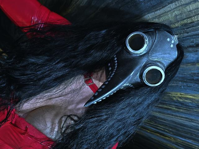 Devasha with black wavy hair wearing a red shirt, a black mask with round eye holes, and a red collar, looking up with a slightly open mouth.
