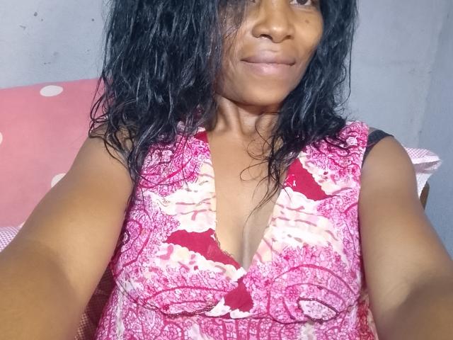 MiaRomia, com cabelo escuro e molhado, pele marrom média e um vestido floral rosa com um decote em V profundo, mostrando seus pequenos seios e um sorriso discreto em uma selfie..