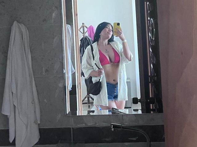 Elizaa, com cabelo preto, usando um top de biquíni rosa que mostra seus seios e shorts jeans, vestindo uma camisa branca aberta, tirando uma selfie no espelho em um banheiro..