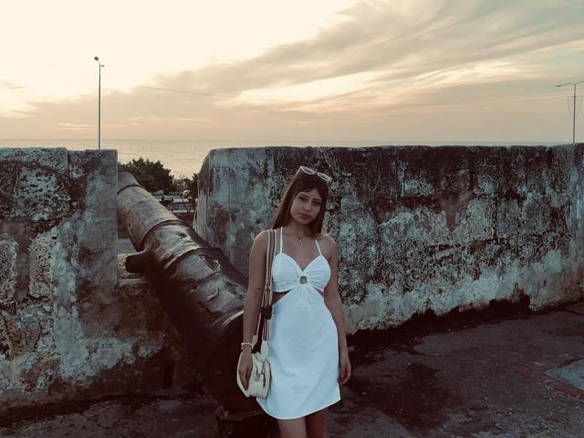 MadissonBlue with small tits and a white dress stands in front of a weathered stone wall with a large pipe, holding a beige handbag, at sunset.