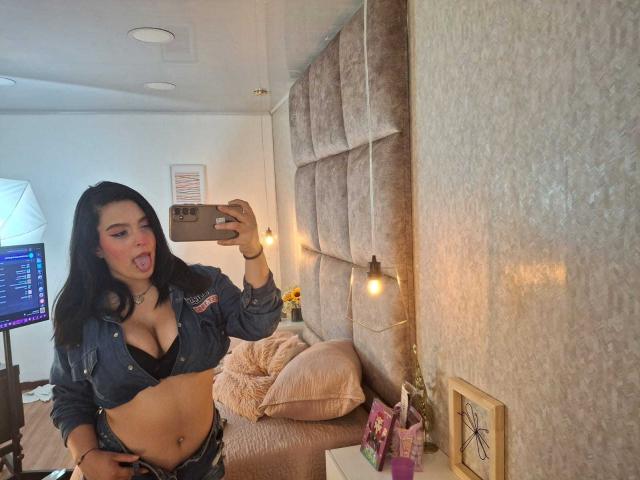 StacyAnzola with black hair, tongue out, wearing an open denim shirt exposing her big tits and black bra, taking a selfie in a bedroom with a tufted headboard.