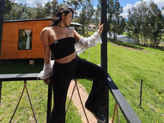 ElizabethMacy with dark hair, wearing a black strapless top, black pants, and white sleeves, leaning on a black railing, showing her tits and ass, outside on a sunny day.