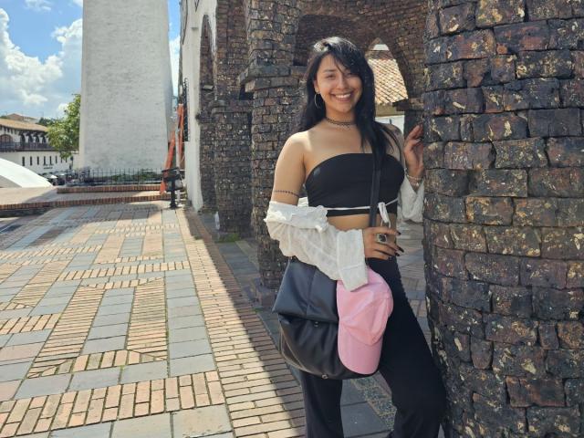 ElizabethMacy with long black hair, smiling, wearing a black strapless top, white off-shoulder shirt, black pants, pink cap, black bag, standing against a brick wall, holding phone.