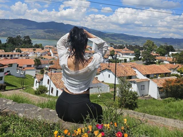 ElizabethMacy, butt visible through a white, long-sleeve, off-shoulder top, kneeling with hands in curly black hair, wearing black pants, overlooking a red-roofed village with a blue sky.