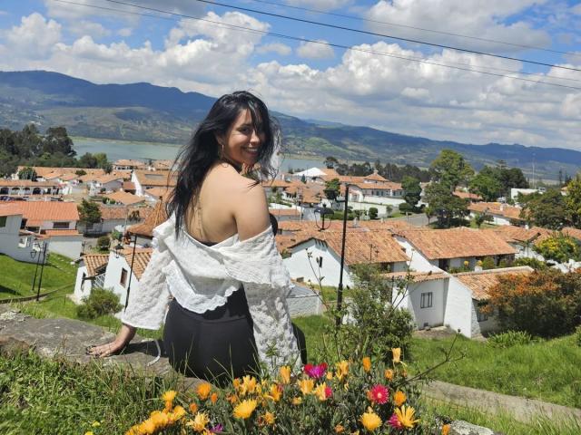 ElizabethMacy with long black hair, off-shoulder white top, and black pants, smiling over a grassy hill, surrounded by yellow flowers, red roofed houses, and a mountainous landscape.