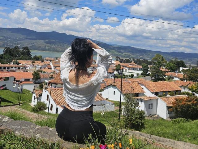 ElizabethMacy with black hair, white sheer top exposing her ass, and black pants, sitting on a stone wall, hands in hair, overlooking a village with red roofs and green hills.