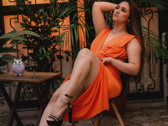 MariahVictoria with orange dress, crossed legs, black heels, and anklet, sitting in chair, arm behind head, green plants, ornate wall, small table with owl and glass.