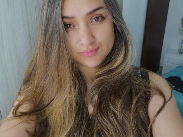 MariahVictoria with long, wavy brown and blonde hair, brown eyes, and pink lips, wearing a black sleeveless top, smiling softly at the camera.