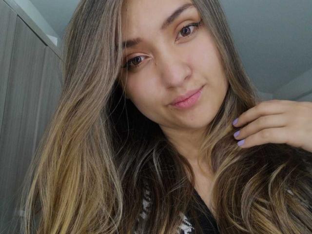 MariahVictoria with long brown hair, subtle makeup, and pink lips, showing off her hand with purple nail polish, smiling softly, tits not visible, wearing a black and white top.