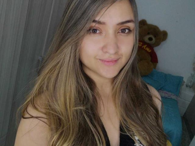 MariahVictoria with long brown hair, blue eyes, and a small smile, wearing a black top, showing her tits. Brown teddy bear in the background. Bedroom setting.