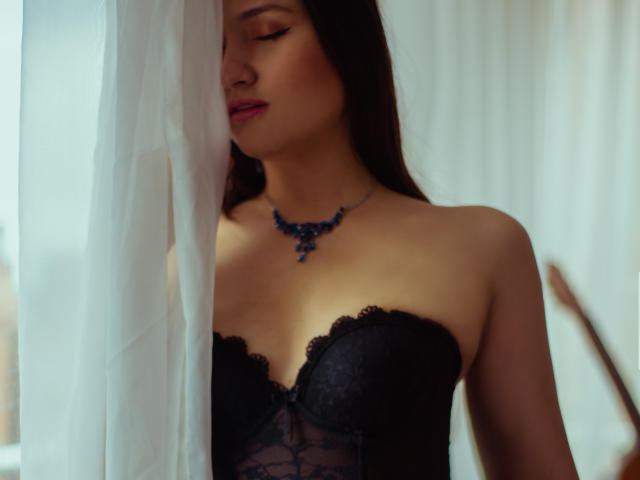 MariahVictoria with long dark hair, closed eyes, black lace strapless corset, blue necklace, standing by white curtain, soft light, slightly blushed, sexy, delicate, subtle, elegant.