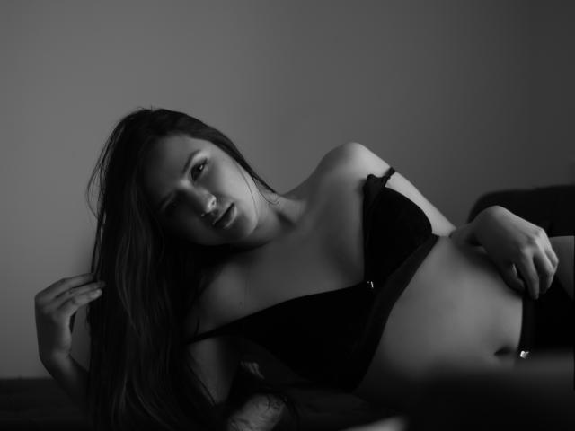 MariahVictoria with long dark hair, black bra, and black underwear, lying on a bed, her hand in her hair, looking seductively at the camera, her tits slightly visible.