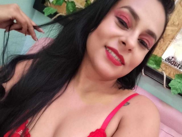 LunaSoler with long black hair, red makeup, red lipstick, red bra, smiling, holding hair, pink chair, small tattoo on right shoulder, medium breasts, light skin.