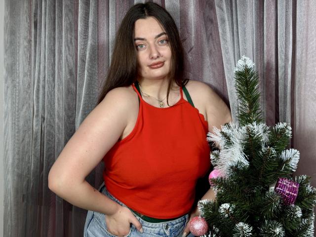 MilanaPrice with long brown hair, red tank top, green bra straps, and jeans, standing next to a small Christmas tree, has a curvy, fair-skinned body, and a cute face.