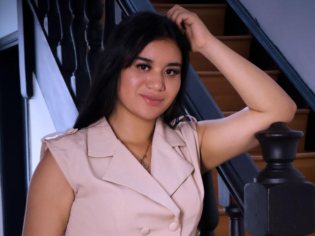 AnieLeeX mit dunklem Haar, heller Haut, kleinen Brüsten, trägt eine rosa, ärmellose Bluse, lächelt, Arm erhoben, Hand im Haar, steht in der Nähe einer Holztreppe mit schwarzen Geländern..