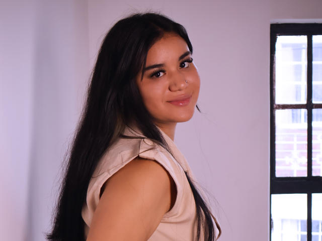 AnieLeeX, com cabelos longos e pretos, olhos castanhos, um pequeno piercing no nariz e um leve sorriso, vestindo uma blusa bege sem mangas, olhando por cima do ombro. Os seios não estão visíveis..