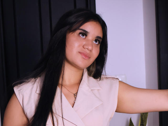 AnieLeeX, con cabello largo y negro, una camisa rosa claro y una sonrisa sutil, mirando ligeramente hacia arriba con el brazo derecho levantado. Tiene ojos marrones, piel clara y una nariz pequeña..