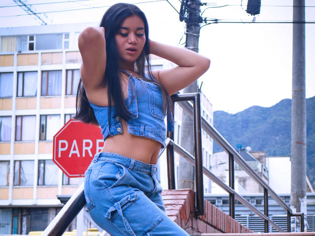AnieLeeX mit langen, schwarzen Haaren, die ein blaues Jeans-Crop-Top und dazu passende, hochgeschnittene Hosen trägt, lehnt an einer Ziegelwand, die Hände im Haar, und zeigt ihren schlanken, gebräunten Bauch..