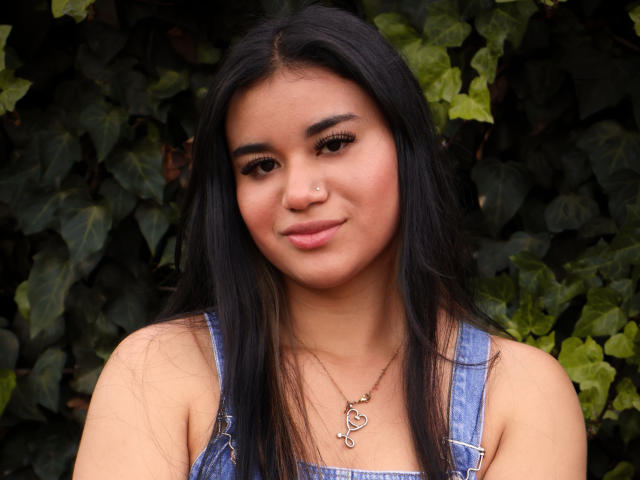 AnieLeeX hat langes, schwarzes Haar, einen Nasenring und trägt Jeansoveralls mit einer Herz-Kette. Sie lächelt leicht, trägt dunklen Eyeliner und hat ein süßes Gesicht..