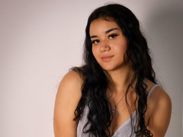 AnieLeeX, con cabello negro largo y ondulado, vestida con una camiseta gris, tiene una sonrisa encantadora, piel bronceada y ojos marrones. Lleva un collar con un pequeño corazón..