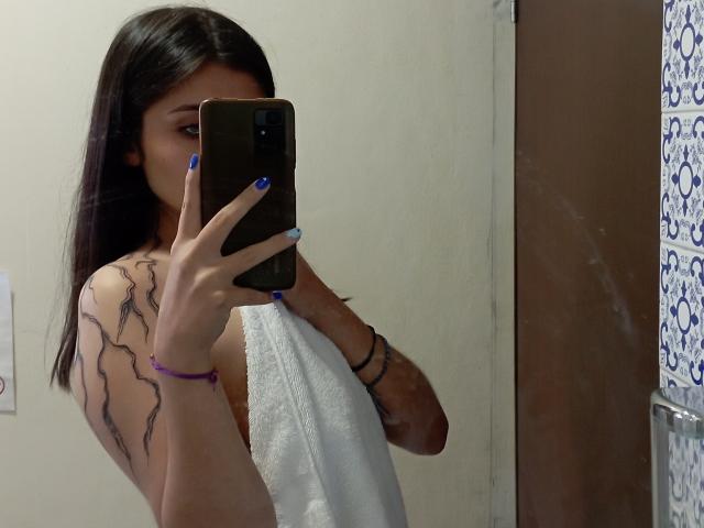 SarahhTanyas con capelli scuri, unghie blu, che tiene un telefono, copre i seni con un asciugamano bianco, con un tatuaggio visibile sul braccio, in piedi nel bagno, selfie allo specchio, braccialetto al polso..