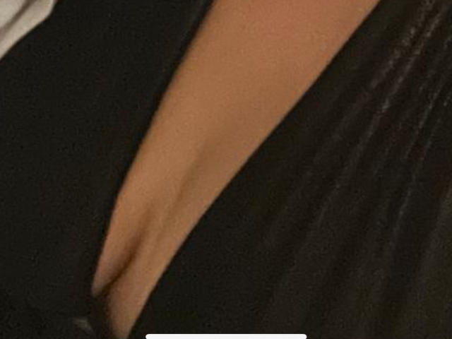 PlaisirHumide's cleavage, showing a deep, black-straped neckline that reveals a tan, smooth, curved, and slightly glossy breast. The image focuses on the upper chest area.