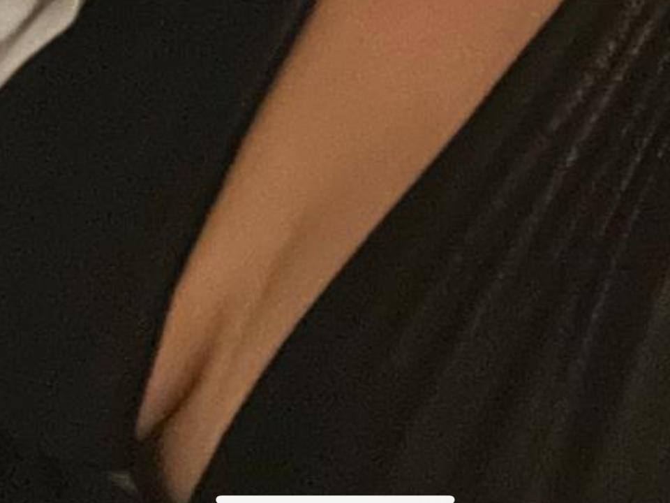 PlaisirHumide's cleavage, showing a deep, black-straped neckline that reveals a tan, smooth, curved, and slightly glossy breast. The image focuses on the upper chest area.