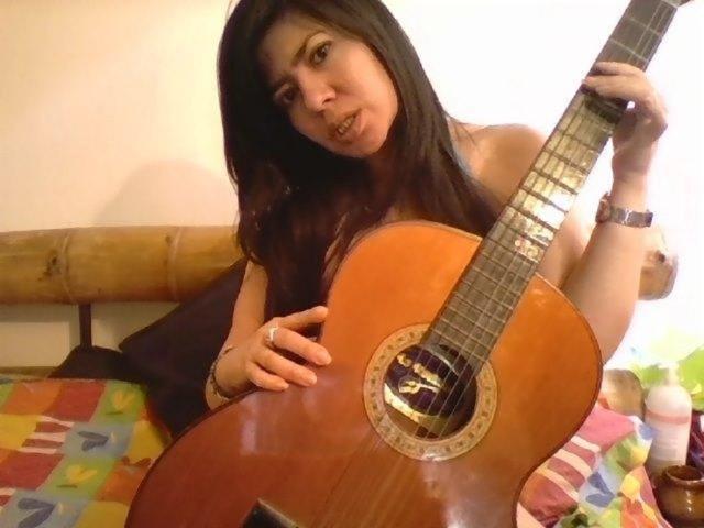 NishaX mit langen, dunklen Haaren, die eine akustische Gitarre aus Holz hält, sitzt auf einem bunten Bett, trägt ein schwarzes Top ohne Ärmel und blickt mit einem leichten Lächeln in die Kamera..